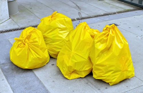 Four large yellow rubbish bags made from durable plastic are tightly knotted at the top, filled with waste materials, and placed on a grey concrete pavement driveway. The bags are positioned in a loose cluster, with some leaning slightly to the side, and are likely awaiting collection or disposal. The surface around them is smooth, with visible concrete slabs and a metal drain grate near the top left corner of the image, suggesting an outdoor urban or residential setting. The lighting appears natural, casting faint shadows beneath the bags, and the scene reflects typical rubbish removal activities in an English town or neighbourhood, aligning with services provided by Business Waste Removal Aldgate. The overall environment is clean, with no other objects or debris nearby, indicating an organized waste collection process.