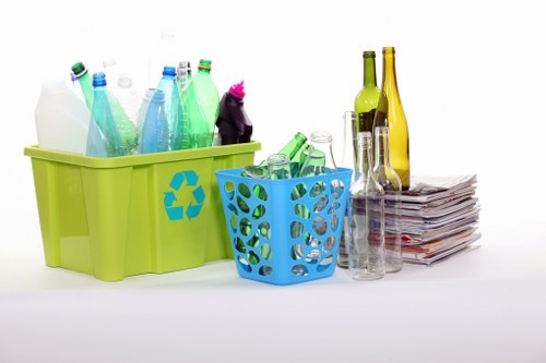 A collection of recyclable household waste materials arranged on a white surface, including a large lime green plastic bin containing various plastic bottles in different shades of blue, green, and translucent, with some bottles having labels and caps. In front of the green bin is a smaller blue plastic basket with a circular cut-out pattern, holding additional plastic bottles, some with caps, and a clear glass container. To the right, there are several empty glass bottles in shades of green and yellow, varying in height, along with a neat stack of crumpled newspapers or paper waste. The background is plain and white, providing a clear view of the waste items. This setup reflects typical rubbish removal and recycling preparation as offered by businesses like Business Waste Removal Aldgate, specialising in rubble and general waste collection in the Aldgate area, with a focus on eco-friendly disposal practices and efficient waste management.