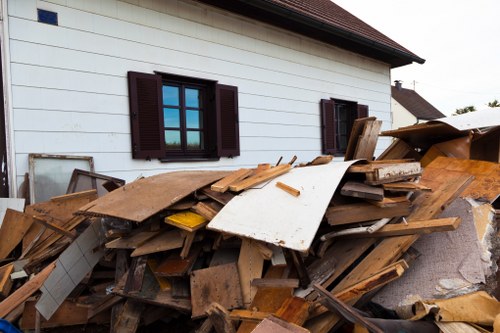 A large pile of mixed construction and domestic waste materials is situated next to the side of a white, wooden-clad building with dark brown window shutters. The pile includes broken wooden planks, panels, and boards of various sizes, some with visible nails and rough edges, as well as pieces of drywall, cardboard, and other debris. The materials are stacked irregularly, leaning against the exterior wall and spilling onto the pavement or ground nearby. The background shows a clear sky and additional rooftops of neighboring houses. The scene suggests a site prepared for rubbish removal services, highlighting the type of bulky waste that Business Waste Removal Aldgate commonly handles in the local area, near Aldgate and the broader London region. The overall environment appears to be a residential or commercial property exterior with overgrown items awaiting collection or disposal, consistent with professional rubbish clearance operations in the area.