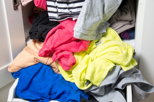A cluttered laundry drawer filled with various pieces of clothing including a bright yellow hoodie, a pink garment, a beige item, a blue fabric, and a grey piece, all appearing crumpled and jumbled together. The surrounding environment suggests an indoor setting, possibly a bedroom or utility area, with visible parts of the drawer and some clothing spilling over the edges. The image highlights the typical mess associated with laundry or clothing management, which a waste removal service like Business Waste Removal Aldgate could assist with by properly clearing and disposing of unwanted textiles and garments in the Aldgate area. The colours are vivid, and the textures range from soft fabric to the structured plastic of the drawer, emphasizing the disorganized state. This scene underscores the importance of professional rubbish and textile removal for domestic or commercial property upkeep, especially within central London’s historic district.