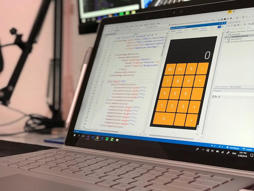 Close-up image of a silver laptop placed on a desk displaying a software development environment with lines of code in a text editor and a large orange calculator interface onscreen, featuring basic a