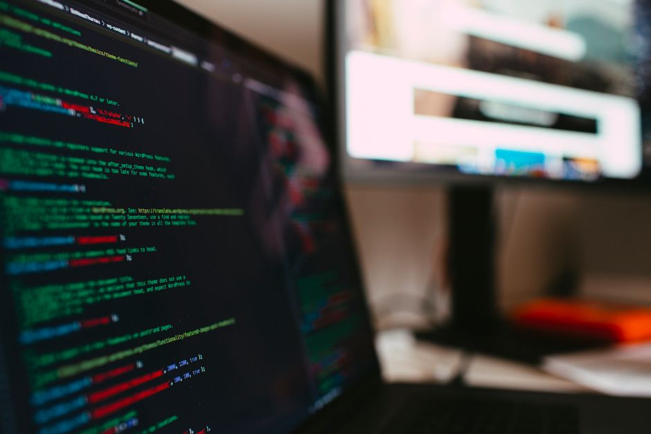 The image displays a dense overlay of computer code in multiple colours, including red, green, blue, yellow, and white, on a dark background. The code consists of various programming syntax elements s