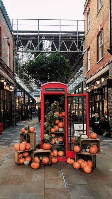 Inside a covered market area with brick buildings on either side, a red vintage-style telephone booth is repurposed as a display for decorative pumpkins and potted plants, forming a seasonal focal poi